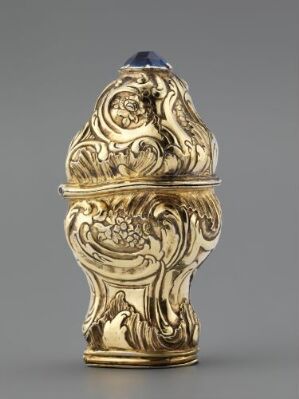  An ornate golden metal object shaped like an elongated egg, featuring intricate Baroque-style relief patterns and topped with a dark-colored dome, set against a neutral gray background.