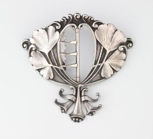  A fan-shaped, silver-colored metal brooch with a stylized leaf design, featuring a reflective vertical central element and ornamental swirls at the bottom against a light gray background. Artist and title are unknown.