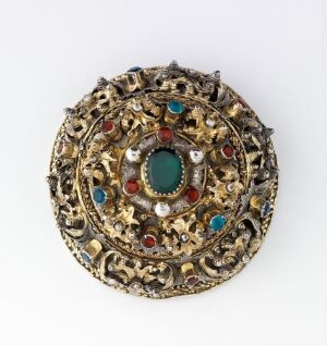  An intricately designed round golden ornament with a central turquoise gemstone, surrounded by concentric circles of red, blue, and clear gemstones set in elaborate gold filigree work on a light grey background.