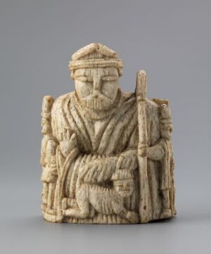  Carved walrus ivory chess piece, "Biskop" (Bishop), with intricate detailing, depicting a seated figure holding a staff and resting a hand on a book, against a gray background. Artist unknown.