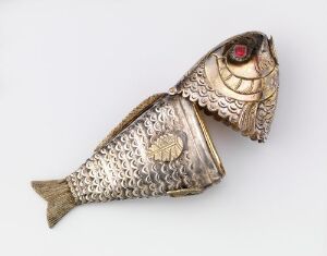  A decorative metallic fish ornament with textured scales in shades of silver, featuring a detailed head with a red gem-like eye and a finely engraved tail, placed against a plain background. The title, artist name, and other specific details are unknown.