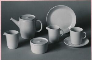  A grayscale photograph of a simple set of ceramic dinnerware, including a teapot, mug, creamer jug, bowl, plate, and saucer, arranged neatly against a gray background.