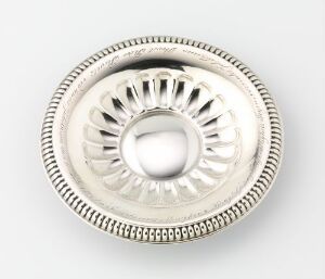  A round, silver metallic object with a convex center and sunburst pattern, featuring beaded inner and twisted rope outer decorative borders against a light background. Artist name and title are unknown.