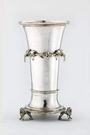  A "Drikkebeger" drinking vessel by an unidentified artist, made of silver with a tapered cylinder shape, ornate gilded garland designs around the rim and base, reflecting a polished silver sheen with warm accents on the decorative elements.