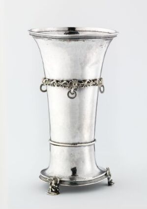  A silver drinking vessel, titled Drikkebeger, from an unidentified artist, with a shiny metallic surface and a chain-like detail encircling its midsection. The vessel stands on small ornamental feet and has a subtly flared rim and base.