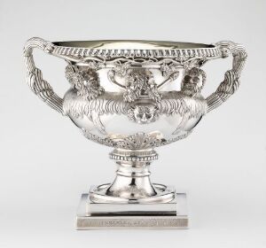  An intricately designed silver trophy with embossed figures and swan-shaped handles on a square base, set against a light grey background. The craftsmanship features classical motifs and detailed embellishments, reflecting light and showcasing artistic skill.