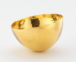  A handcrafted bowl made of hammered gold by artist Hedvig Glomsrød Sommerfeldt, featuring a reflective interior with tarnished spots and a matte exterior. The bowl is placed against a neutral background, emphasizing the metal's golden hue.