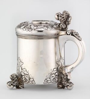  A silver drinking jug ("Drikkekanne") by Mikkel Jensen with ornate baroque style engravings including botanical motifs on the base and lid, and featuring a coin in the lid. The jug reflects light off its polished, cylindrical silver body and is supported by decorative scrolled feet, while the handle is clear and elegantly curved.