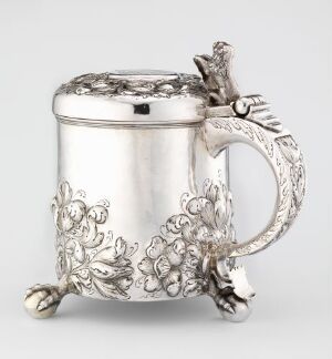  A polished silver drinking vessel "Drikkekanne" by Herman Brouer with a cylindrical body and a richly detailed handle featuring vine and floral motifs. The jug displays smooth surfaces with a hinged lid topped by a decorative finial reflecting meticulous silversmith techniques, with a balance of bright and matte finishes enhancing the ornate design.