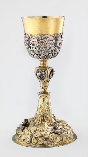  An ornate golden goblet with a polished gold interior and detailed silver floral patterns on the exterior, featuring a decorative stem and a broad, intricately designed base with sculpted figures and patterns.