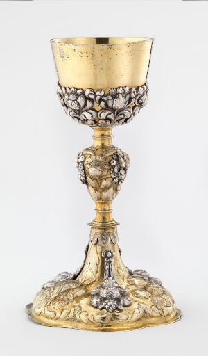  An elaborate golden chalice with a smooth cup and ornately decorated stem and base featuring grapevine motifs and possibly floral or figural embellishments, set against a plain, light background. Artistname and title are unknown.