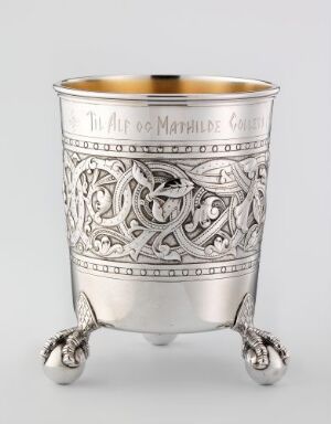  An ornate silver or pewter cup with intricate embossed patterns featuring interlacing lines and stylized animals, standing on three clawed feet, with a notionally gilded interior visible at the rim.