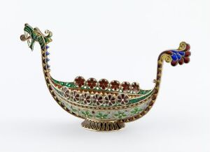  An ornate, jewel-encrusted vessel resembling a bird-boat hybrid with a peacock feather motif on the tail, intricate green and red stonework decorating the hull, and delicate filigree at the base, set against a white background.