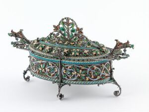  An ornate oval-shaped metal container with filigree and colored inlay work on a filigreed base with four decorative legs. The lid features a large, detailed finial and the sides have scroll or creature-shaped protrusions. The container is predominantly silver with green, white, and red accents.