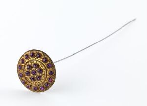  An ornate round brooch with a gold-tone setting and concentric circles of purple gemstones on a white background, with a thin, straight pin extending diagonally from its edge.