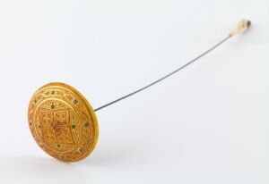  A golden circular spinning rattle with intricate designs, connected by a string to a light-colored wooden handle against a white background. Artist name and title are unknown.