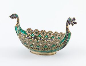  A decorative ornate boat-like object with a bird-like design at the ends, featuring a rich mosaic of green, red, blue, and white colors accented with gold details against a neutral background.