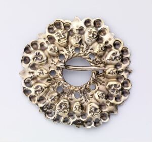  An ornate circular metallic object resembling a brooch with a central hole, adorned with multiple embossed faces showcasing varied expressions, and finished in a gold or brass color.