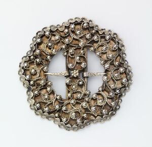  A circular silver-grey metallic brooch with intricate swirl patterns resembling stylized flowers or shells, featuring a cross-shaped negative space at its center, set against a white background. Artistname and Title are unknown.