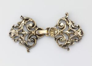  An ornate, symmetrical golden decorative object, possibly a clasp or brooch, with Baroque or Rococo style motifs including scrolls and leaf patterns, showcased against a light grey background.