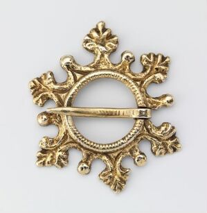  An ornate, gold-colored brooch with a circular center and fleur-de-lis extensions on a light grey background, intricately designed with a polished finish and a visible fastening pin on the back.