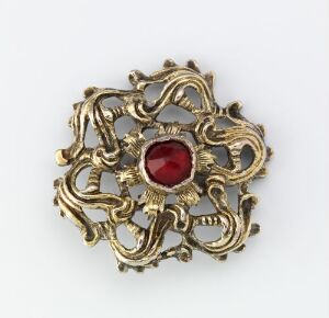  An antique gold-tone brooch with intricate filigree metalwork surrounding a deep red faceted gemstone, set against a plain light grey background.