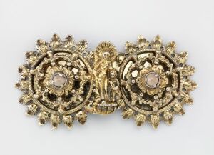  An intricately designed gold brooch with two symmetrical circular motifs, each featuring a large central gem surrounded by radial smaller gems, with a raised human figurine at the center, all set against a light grey background. Artist name and title are unknown.