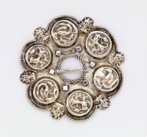  An ornate silver brooch with interconnected circular elements featuring embossed animal or mythical figures, showcasing a lustrous finish with a vintage patina set against a neutral grey background. Artist name and title are listed as unknown.