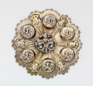  An antique circular gold-toned brooch with a central gem-like focal point, surrounded by eight shield-like crests with raised heraldic designs, set against a neutral background. Artistname and title are unknown.