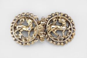  A pair of antique-looking golden ornate clasps or buttons, each with an intricately designed circular frame and a central motif of a stylized heraldic animal, against a light, neutral background.