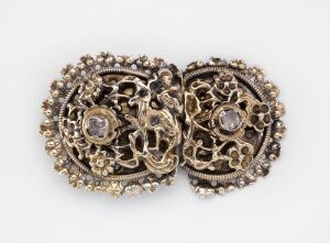  A pair of vintage, symmetrical jewelry pieces with a darkened metal base and ornate floral openwork design, featuring central lighter-colored stones or cameos and numerous small sparkling elements accentuating the intricate patterns.