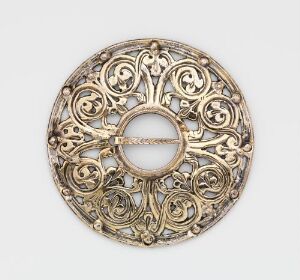  A decorative circular gold brooch with intricate scrollwork, possibly of Celtic or medieval inspiration, against a neutral background.