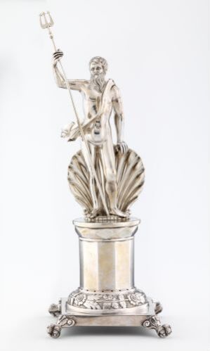  A slender, silver-toned metallic sculpture of a classical male figure standing with a raised right arm holding a staff, draped in traditional garments on a decorative pedestal with a plain, light background. Artist name and title are unknown.