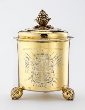  An ornate golden container with lion paw feet and an elaborate embossed coat of arms flanked by wings on its surface, topped with a finial resembling an artichoke or pine cone on the lid.