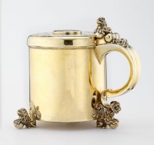  A gilded silver drinking jug titled "Drikkekanne" by Romanus Fredriksen d.e. Møller, with a cylindrical body, coin detail, and ornate decorative elements on the handle and base. The jug has a gleaming golden appearance and showcases baroque-inspired motifs.