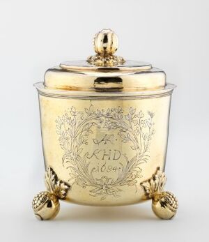  A highly polished golden decorative container with an engraved crest and foliage patterns, standing on three spherical feet with a rounded finial on the lid, set against a neutral background. Artist name and title are unknown.