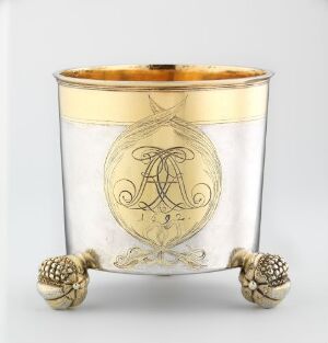  A small, vintage glass container with smoky transparency and a golden metal band featuring an ornate 'A' and 'T' monogram along with the year '1913', capped with a gold rim and supported by intricately designed gold-tone feet.