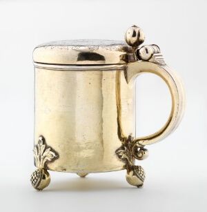  An antique, silver-toned metal mug with a curved handle featuring ornate foliage designs, and decorative elements near the base resembling small floral clusters, set against a neutral off-white background.