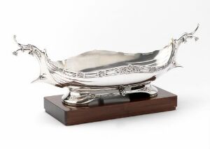  A polished silver gravy boat with ornate details resembling leaves or swirls on both ends, placed on a small rectangular wooden base against an off-white background, exuding a classic and elegant charm. Artistname and title are unknown.