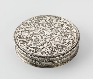  A circular, finely detailed silver box with intricate floral motifs etched into its top and sides, set against a plain, light-colored background. The artist name and title of the piece are unknown.