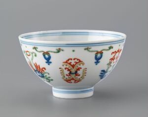  A historical porcelain bowl with bright cobalt blue, red, green, and yellow hand-painted floral and phoenix-like designs, against a light grey background. Artistname and title are unknown.