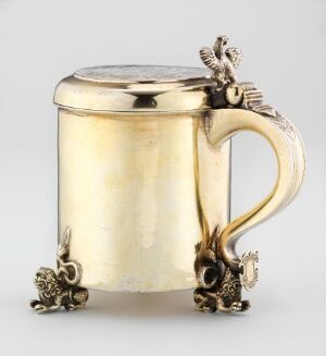  A cylindrical, polished gold-toned tankard with ornate, darker metal accents on the lid thumb-piece, handle, and feet, set against a neutral gray background. Artist name and title remain unknown.