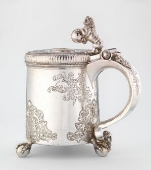  An ornate silver tankard with embossed scrollwork and floral patterns, featuring three spherical feet and a decorative handle with a sculpted thumb-piece on the lid, set against a light gray background.