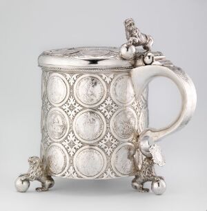  An antique silver tankard with a cylindrical body, embossed medallions, and a sculpted figure on the lid. The tankard stands on three small animal or cherub-shaped feet and features an elegant curving handle.