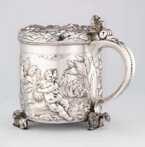  A Baroque-style silver drinking jug, titled "Drikkekanne" by Berendt Johansen, with engraved details depicting cherubs and foliage, highlighted by gilded accents, perched on ornate feet.