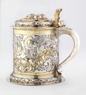  An ornately designed silver or pewter tankard with raised floral and scroll patterns, featuring a curved handle and a decorative lid with a finial. The tankard's design suggests a baroque or rococo style, and it sits against a plain, light background.