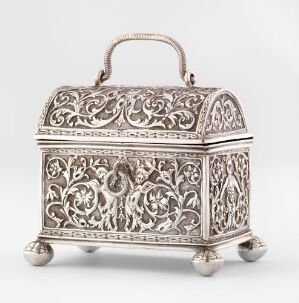  An intricately designed silver-toned metal casket with elaborate scrollwork patterns and a handle on top, standing on four spherical feet against a light grey background.