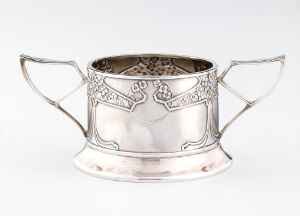  An ornate silver sugar bowl with decorative engravings and handles, set against a light grey background, showcasing its reflective surface and detailed craftsmanship.