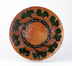  A circular ceramic plate featuring a central terracotta-colored depression with grooved lines and a surrounding rim decorated with raised, stylized green leaves against an orange background, showcasing an earthy, artisanal design.