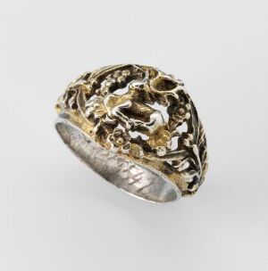  A textured silver ring with gold-tone highlights featuring an organic, lace-like design, displayed against a neutral background. Artistname and title are unknown.